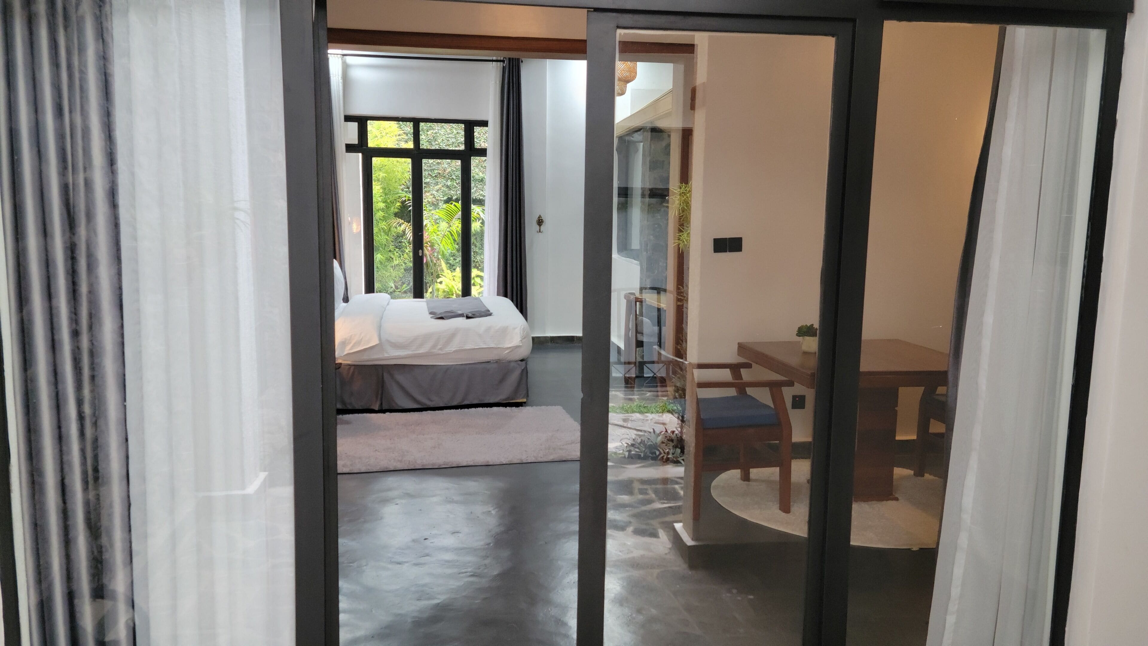 A bedroom with a bed, a small table, and a chair. There is a window with curtains and a view of greenery outside.