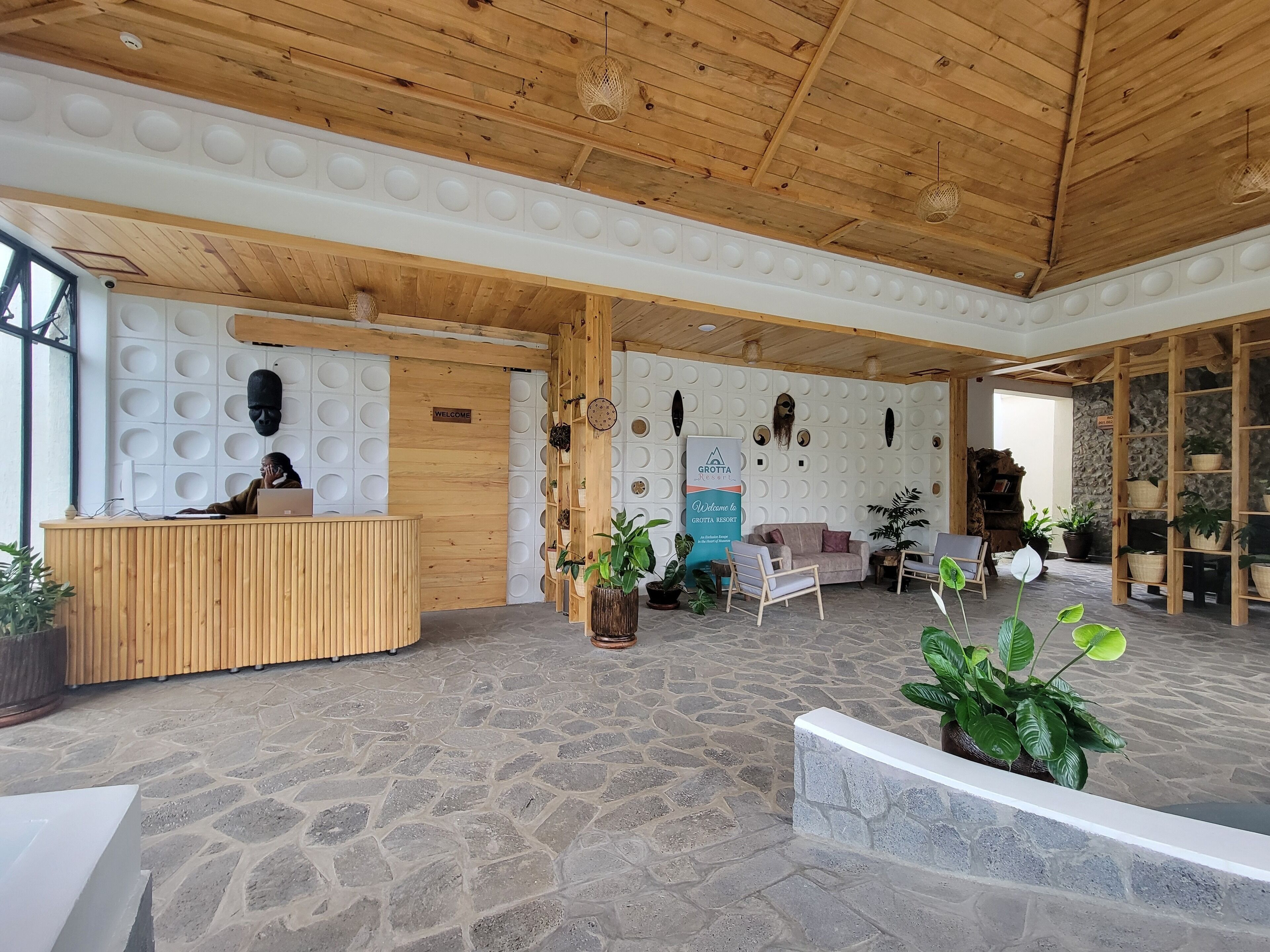 A reception area with a wooden desk, a 'Welcome' sign, and a 'Grotta Resort' sign. There are potted plants and a stone-tiled floor.