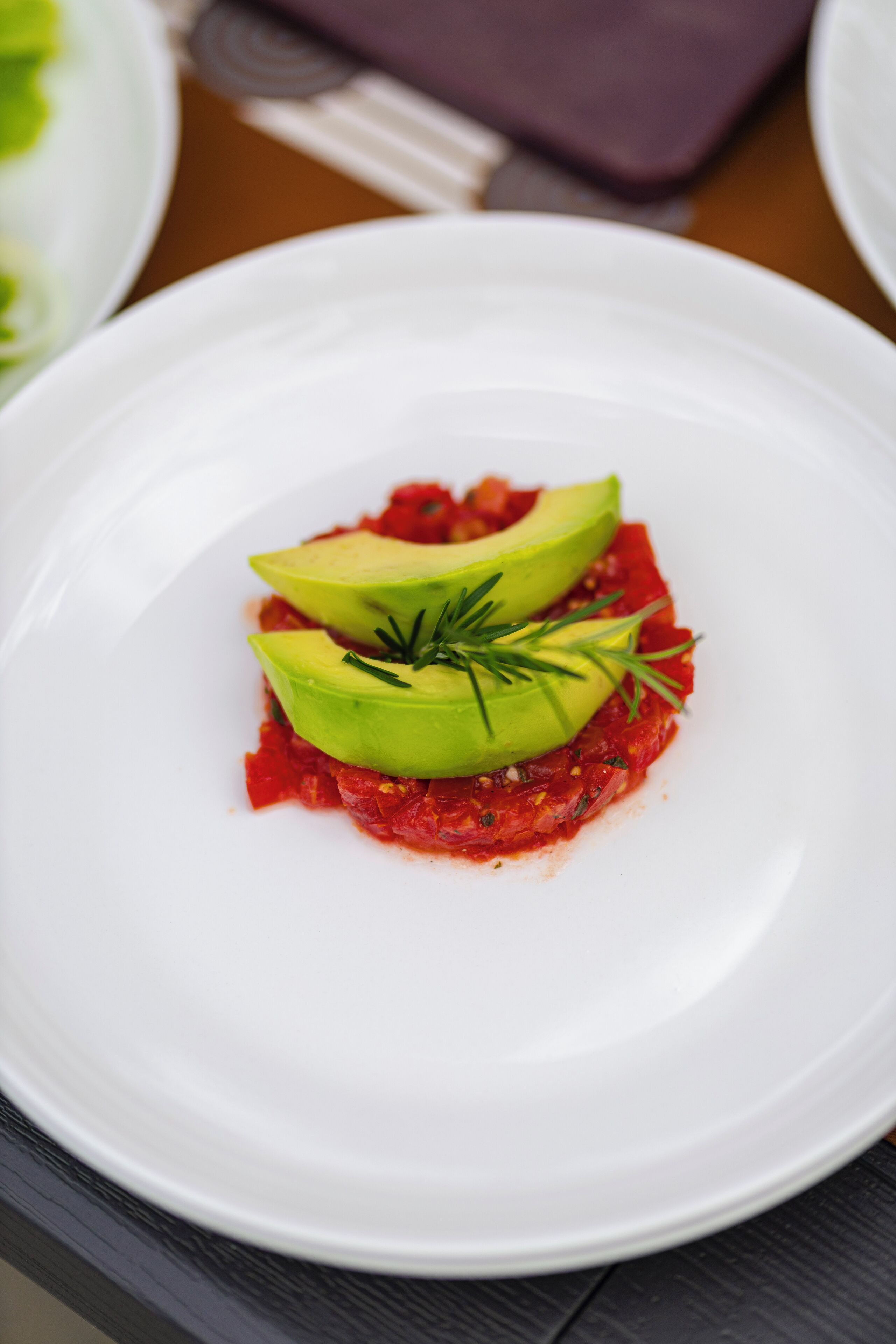Ein weißer Teller mit einer Schale rote Soße, belegt mit Avocadowürfeln und einem Rosmarinzweig.