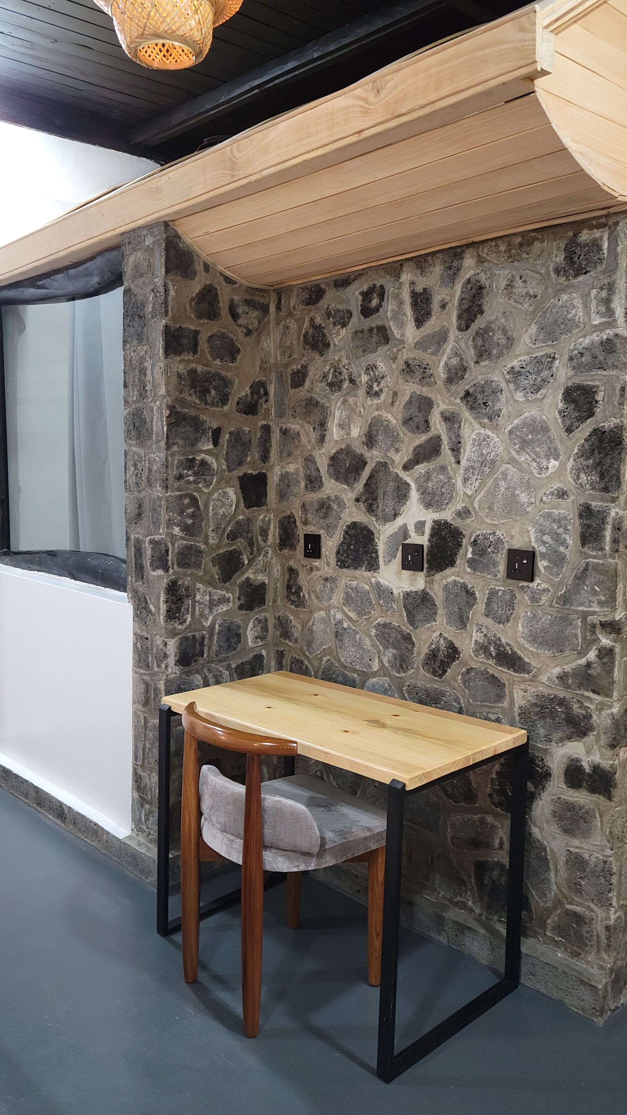 Una pequeña mesa de madera con una silla acolchada en un rincón, junto a una pared de piedra y una ventana.