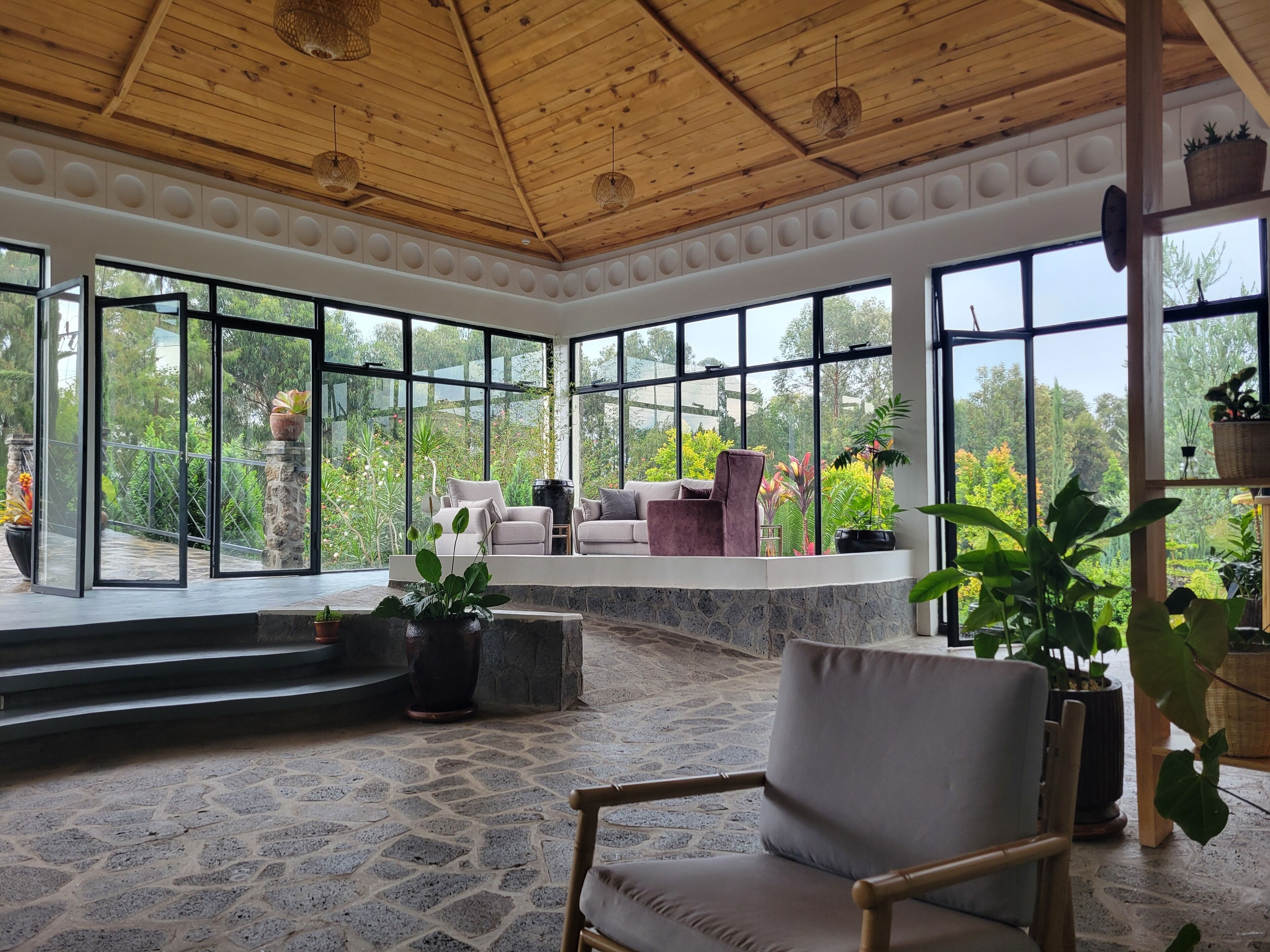 Una habitación amplia con suelo de piedra, ventanas grandes que dan a zonas verdes y techo de madera.