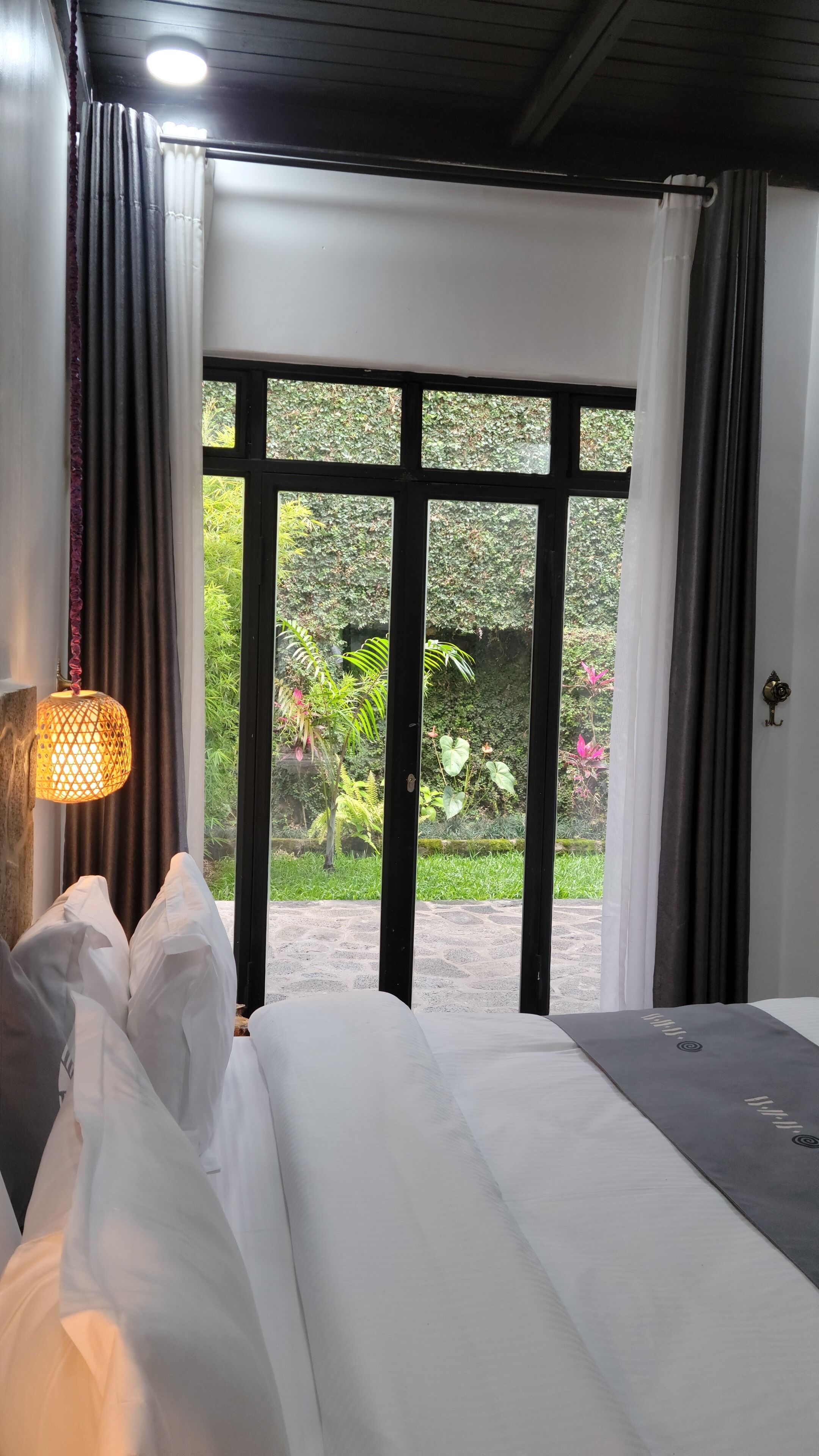 A hotel room with a bed, a hanging lamp, and a view of a garden through large windows.