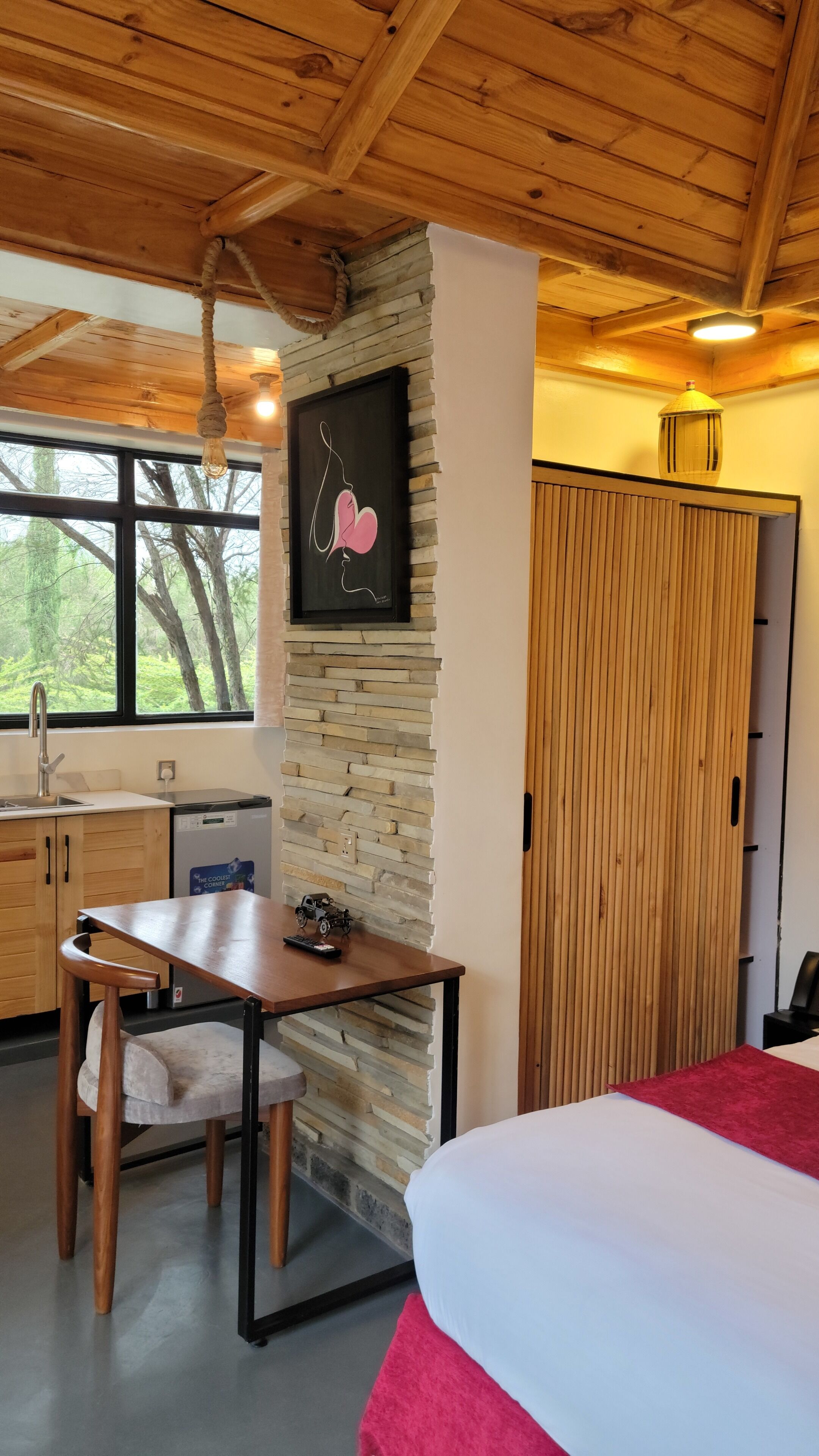 A room with a wooden ceiling, a stone wall, a bed with a red blanket, a table with a toy car, a sink, and a window with a view of trees.