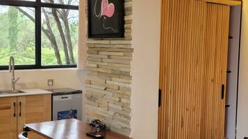 Una habitación con techo de madera, pared de piedra, cama con manta roja, mesa con un coche de juguete, lavamanos y ventana con vista a árboles.