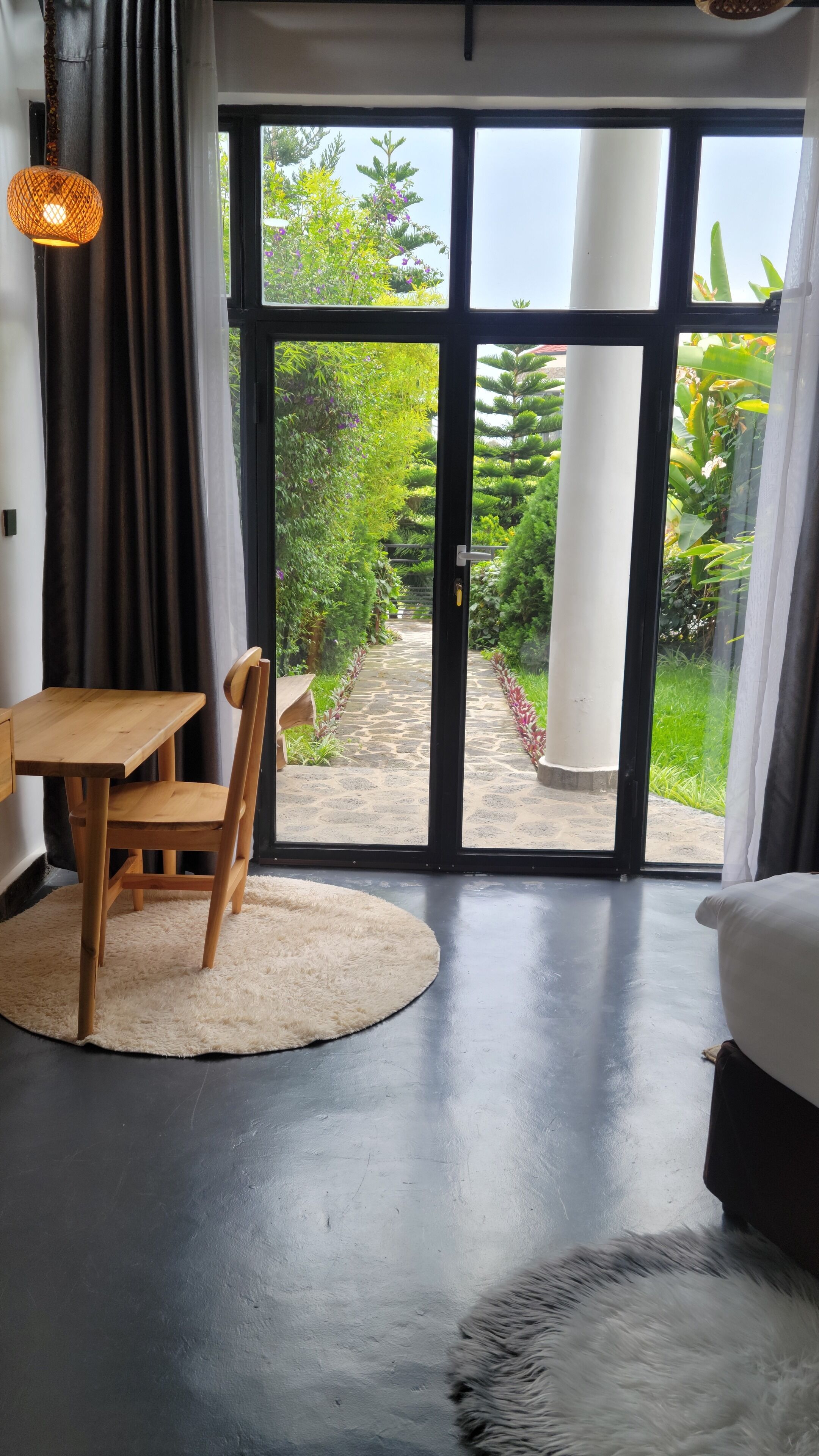 Una habitación con una mesa y una silla de madera, un tapete blanco y un amplio ventanal corredero que da a un jardín.