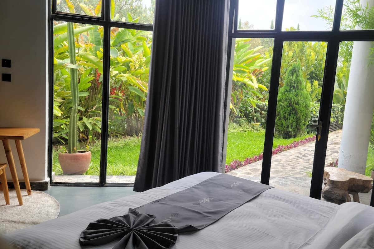 A bedroom with a large window, a bed with a neatly arranged pillow, a wooden side table, and a potted plant.