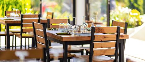 Un restaurante con mesas y sillas de madera, vasos transparentes y vistas a zonas verdes.
