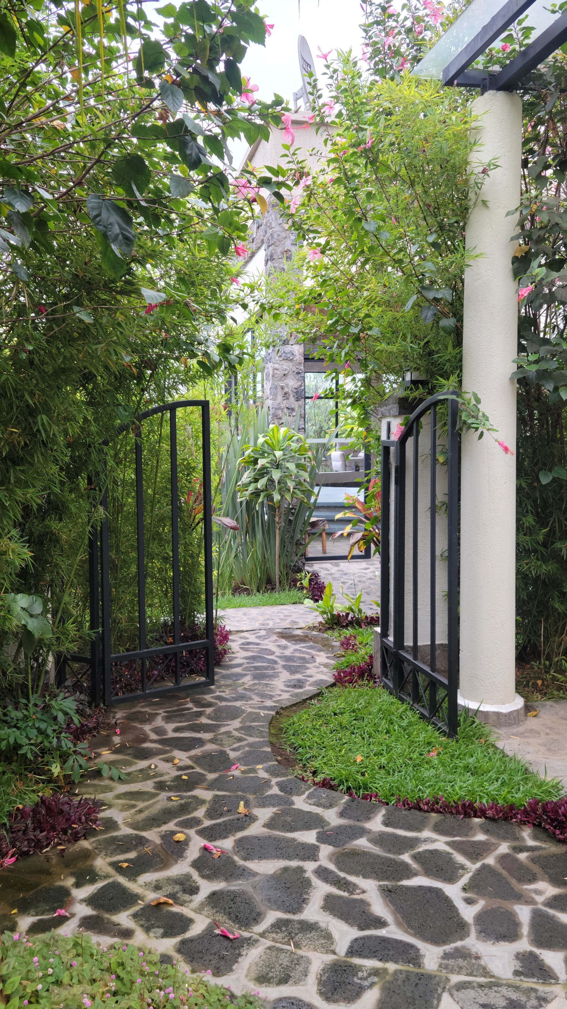 A stone pathway leading to a building entrance, surrounded by lush greenery and a metal gate.