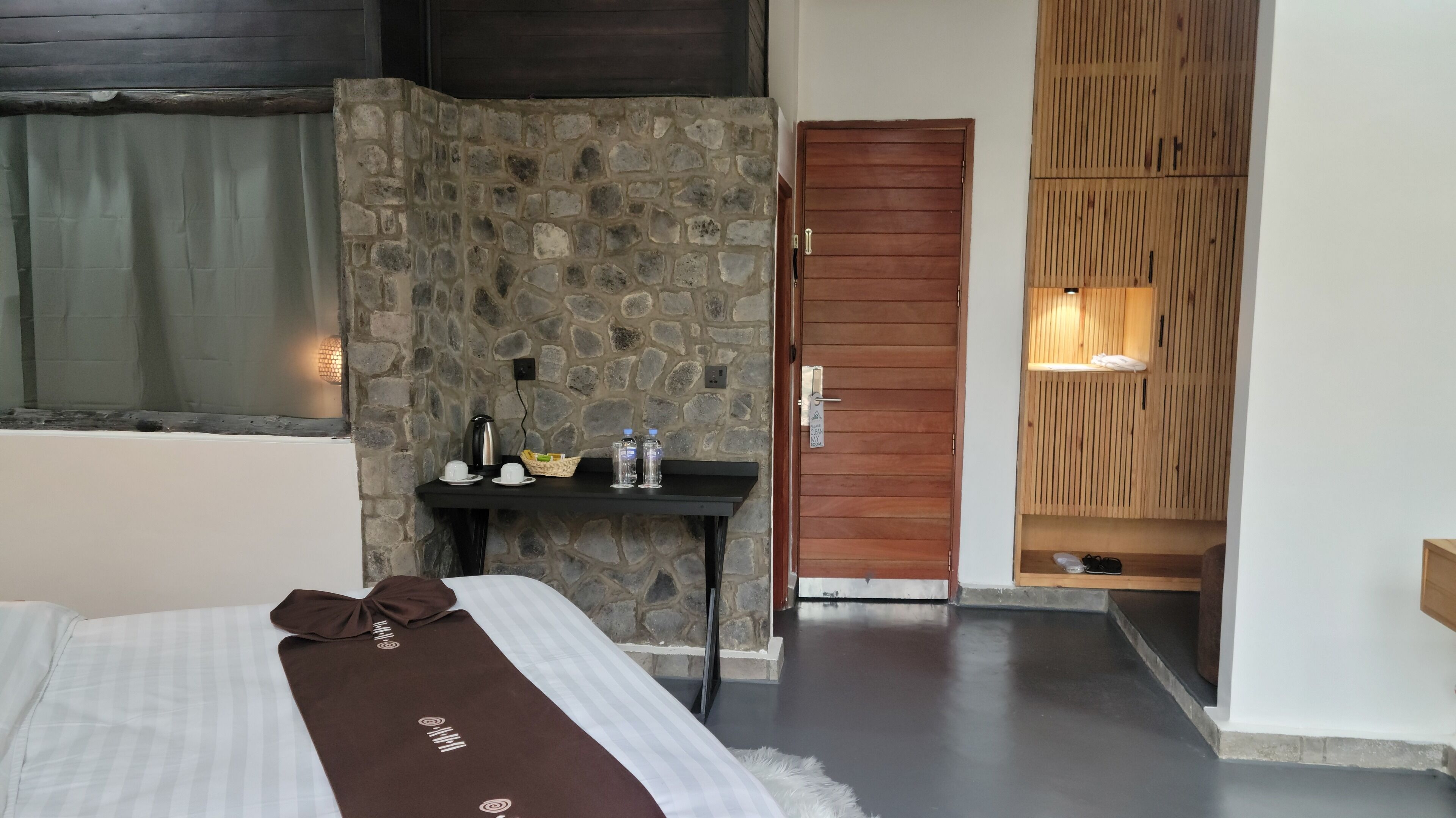 A hotel room with a stone wall, a wooden door, a small table with a kettle and cups, and a bed with a towel and a brown headband.