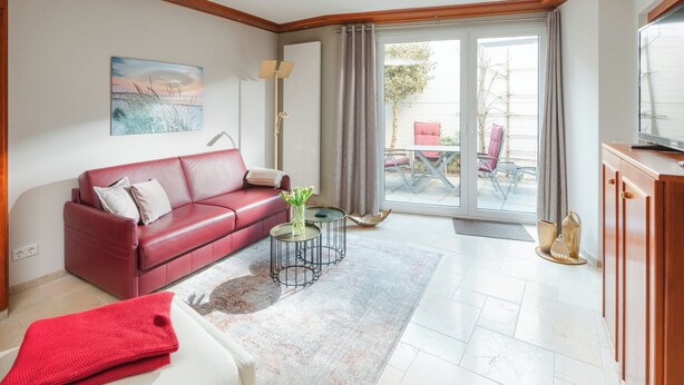 Un salon avec un canapé en cuir rouge, une table basse en verre, un tableau accroché au mur et, par une porte vitrée, une vue sur un patio extérieur.