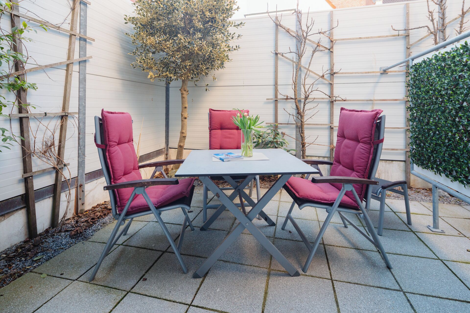 Une terrasse avec une table et deux chaises, entourée d’un mur blanc et d’une haie.