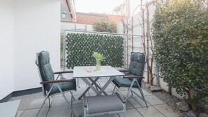 An outdoor patio with a table and four chairs, a small vase with flowers, and a green hedge in the background. - // VACATION APARTMENT BARKE 03 // (Norderney)