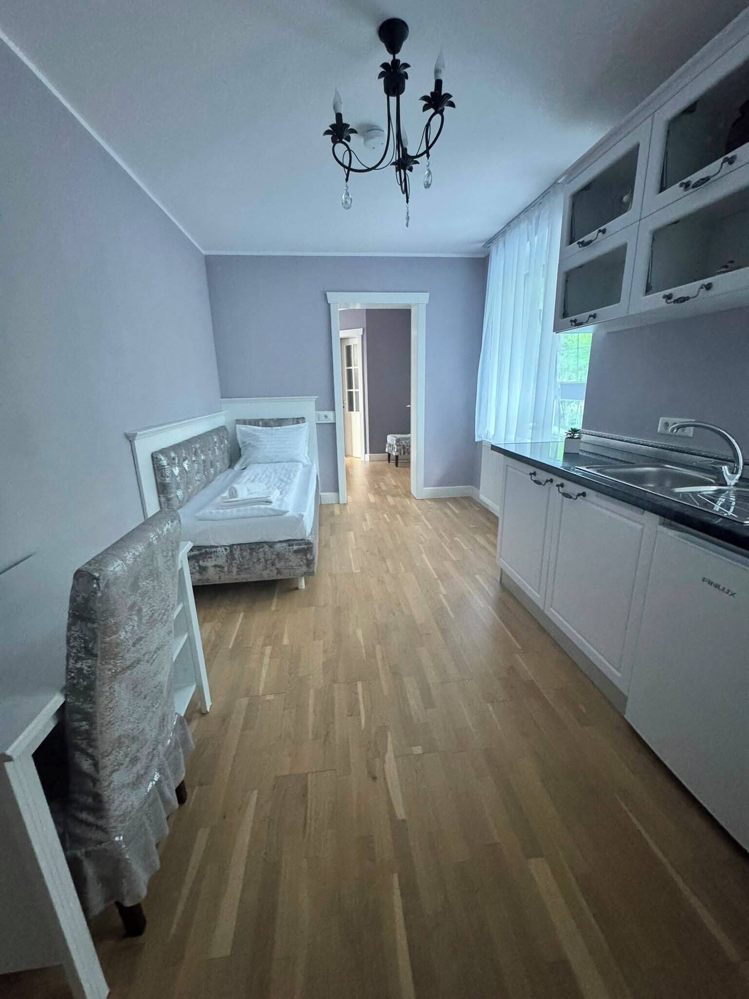 A room with a bed, a chair, a kitchen area, and a hallway leading to another room.