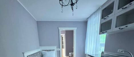 A room with a bed, a chair, a kitchen area, and a hallway leading to another room.