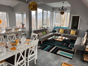 A modern living room with a grey sectional sofa, a dining area with white chairs, and large windows offering a view of trees. - Premont Guesthouse Bran (Bran)