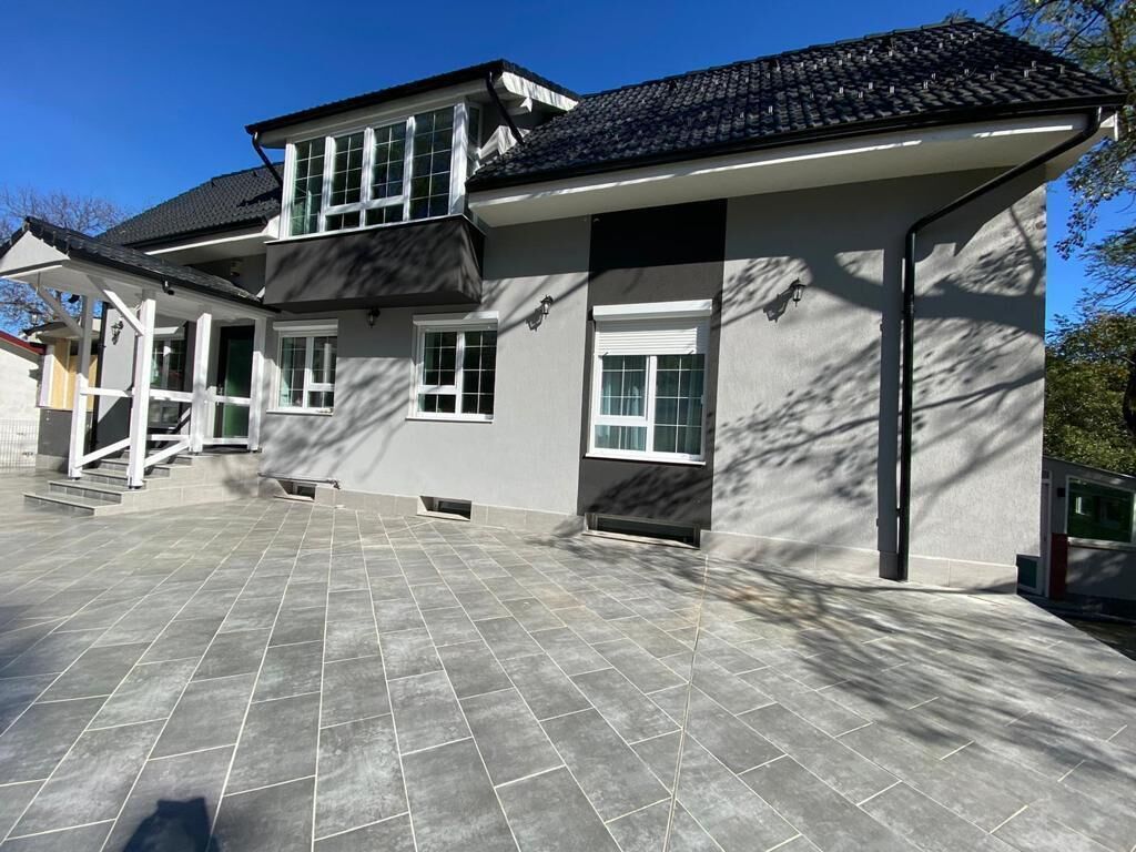 A two-story house with a tiled patio, large windows, and a dark roof.