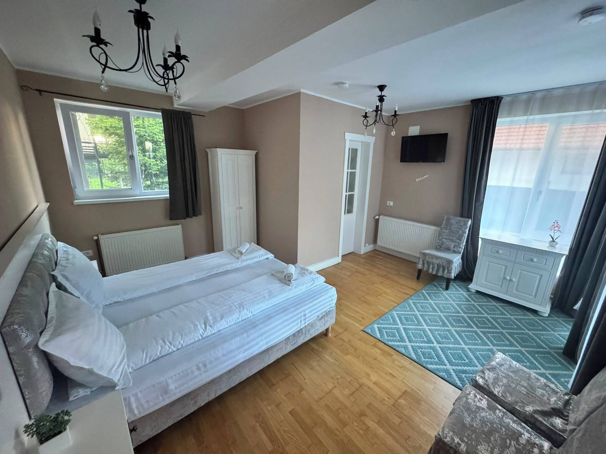 Una habitación de hotel con una cama doble, televisión, radiador, ventana con cortinas, puerta y una pequeña mesa con una silla.