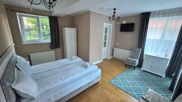 Una habitación de hotel con una cama doble, televisión, radiador, ventana con cortinas, puerta y una pequeña mesa con una silla.