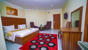 Una habitació d'hotel amb un llit, una taula de treball, dues cadires, una catifa vermella amb dibuixos i un quadre a la paret.