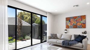 A modern living room with a glass coffee table, a sofa with patterned cushions, and a large abstract painting on the wall. - Townhouse on Tod Glenelg North (Glenelg North)