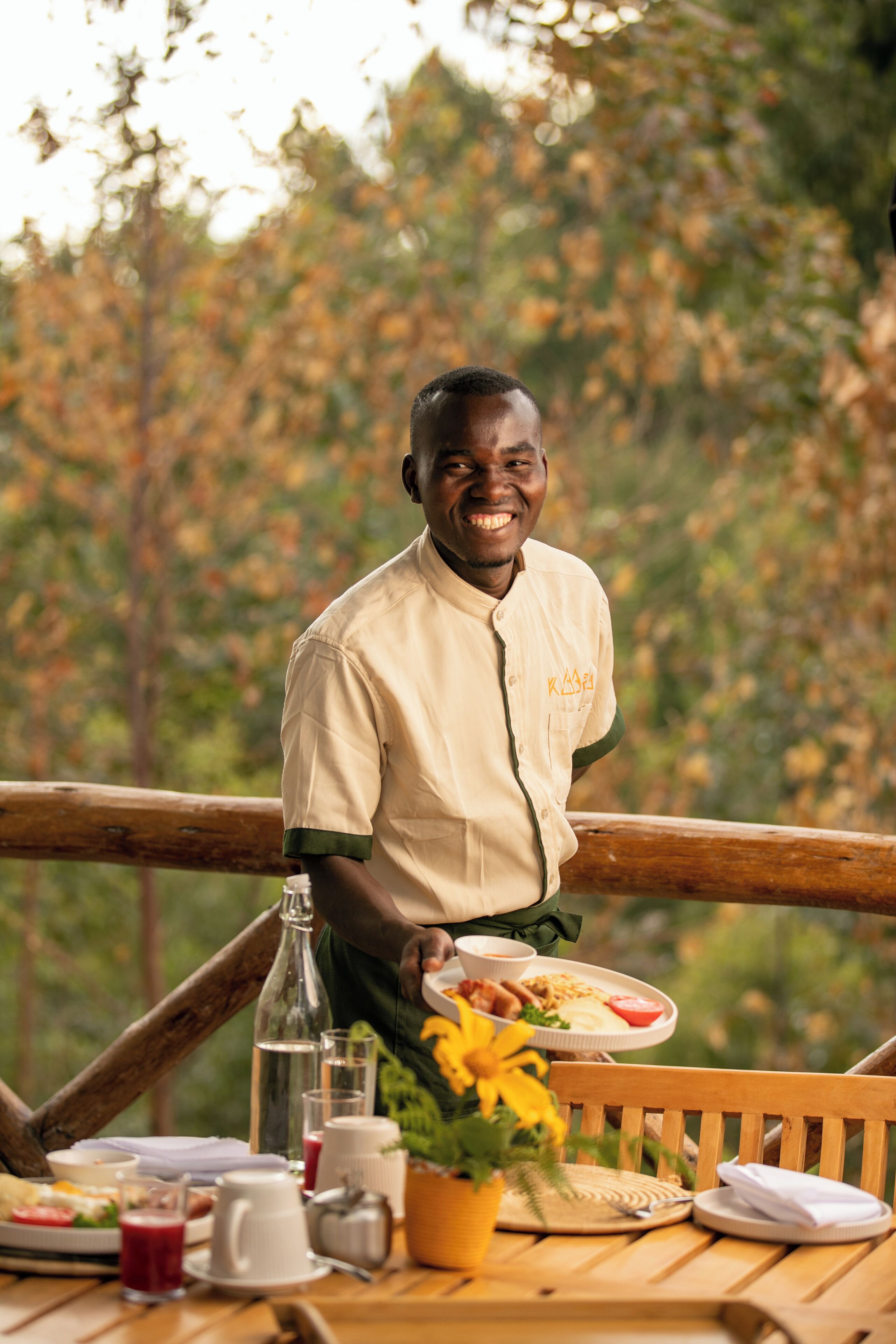 A person serving food on a wooden deck with a table set for a meal, surrounded by autumn foliage.