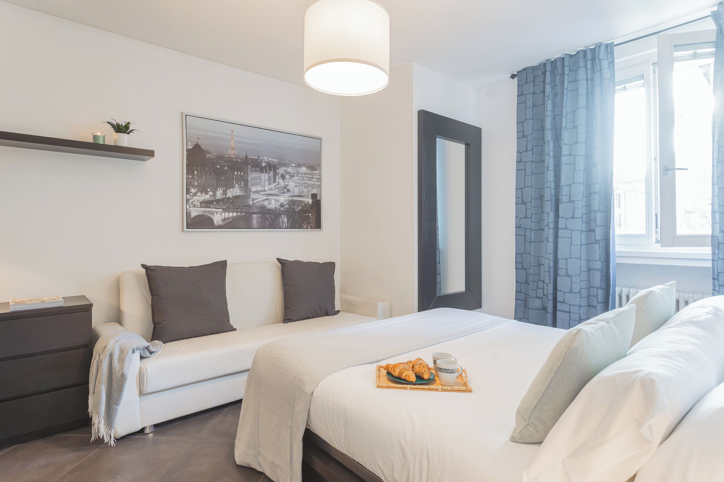 Habitación de hotel con cama, sofá, mesita de noche, lámpara, ventana con cortinas e imagen enmarcadas en la pared.