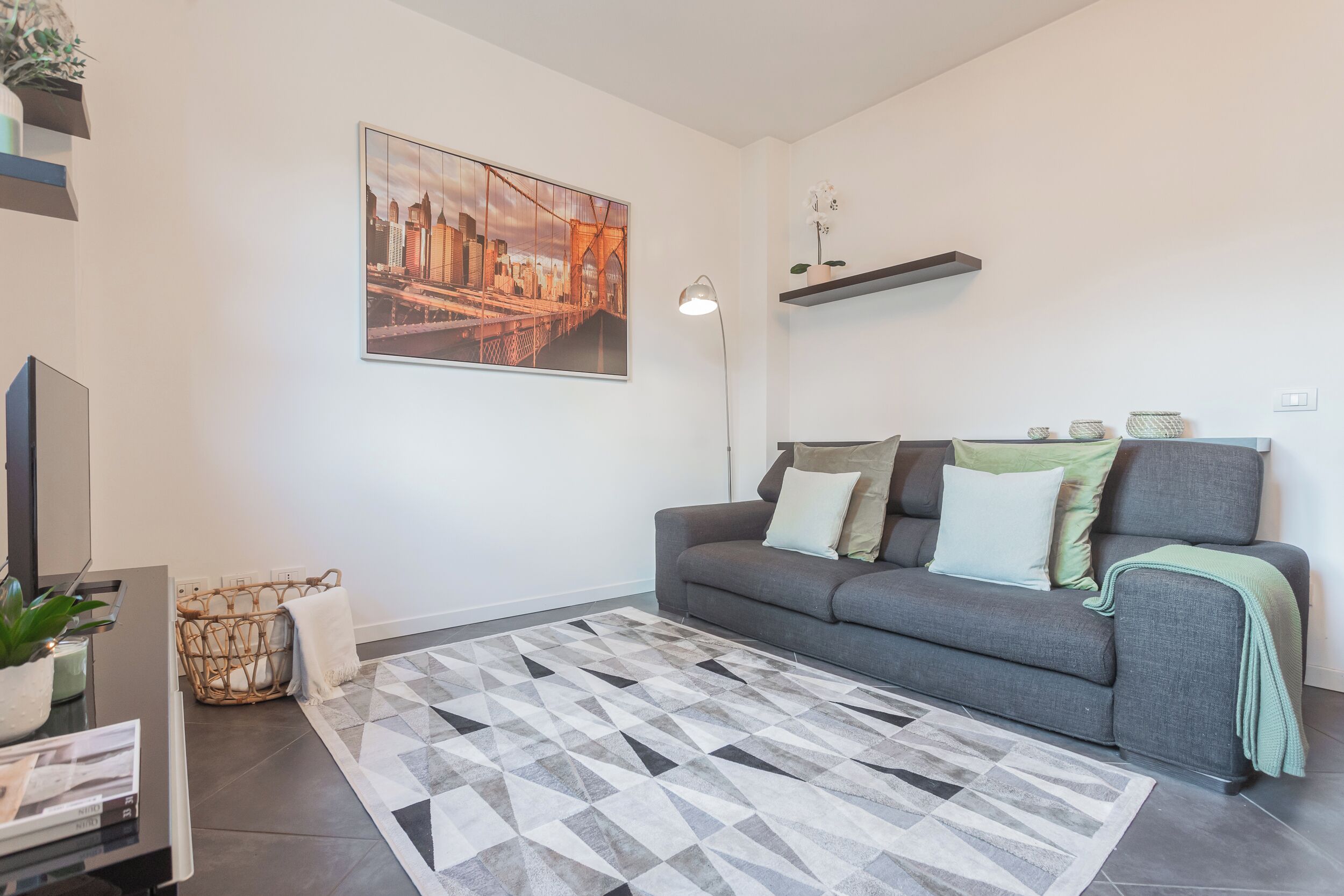 Una sala de estar moderna con un sofá gris, un tapete de estampado geométrico, una pintura de un paisaje urbano y una planta pequeña en una mesita auxiliar.