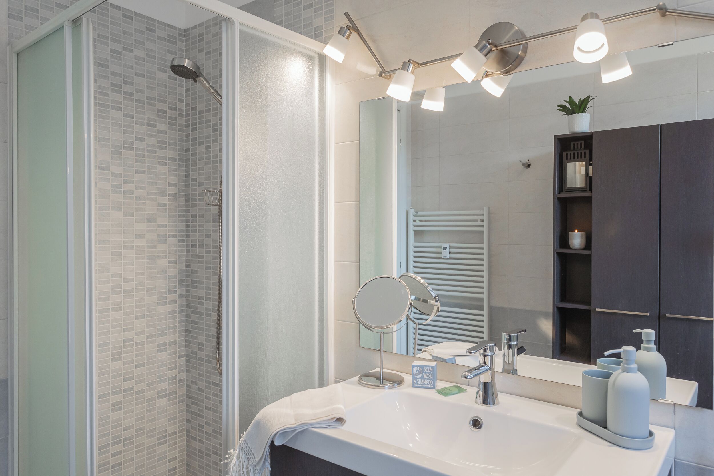 Une salle de bain moderne avec une douche à l’italienne en verre, un grand miroir, un lavabo avec des articles de toilette et un meuble aux tons foncés.