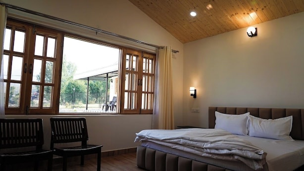 Un dormitorio con cama, dos sillas, ventana con vistas y una lámpara de pared.