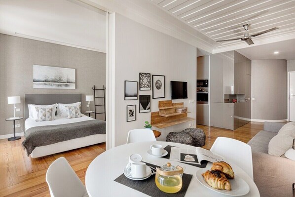 Ett modernt hotellrum med en säng, ett matbord med frukost, en TV och ett litet köksutrymme.