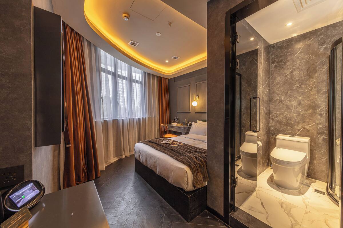 A modern hotel room with a bed, a desk, a bathroom with a shower, and a large window with curtains.