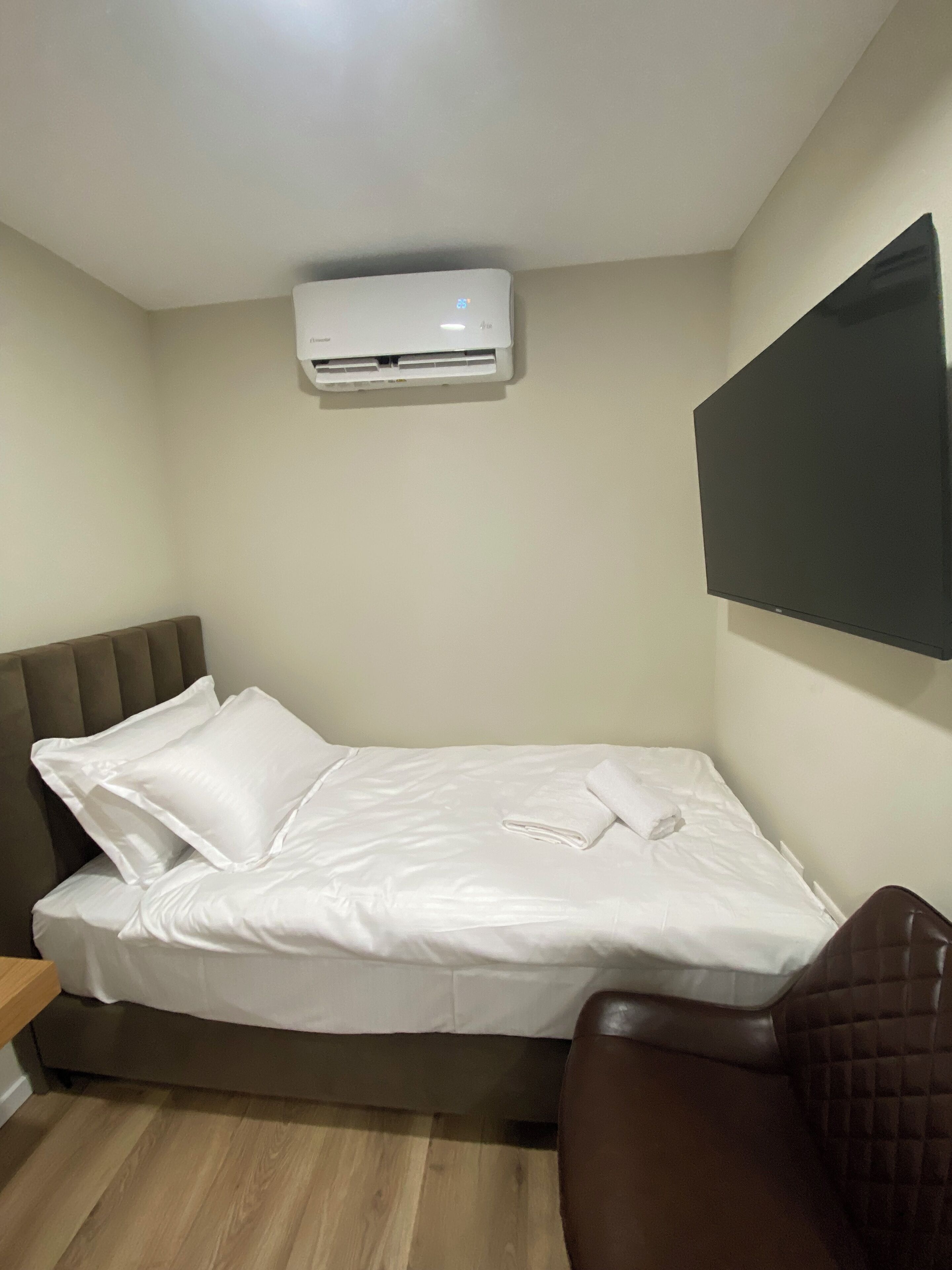 Un lit bien fait, avec du linge de lit blanc, une tête de lit marron, un fauteuil en cuir marron et un téléviseur à écran plat fixé au mur.