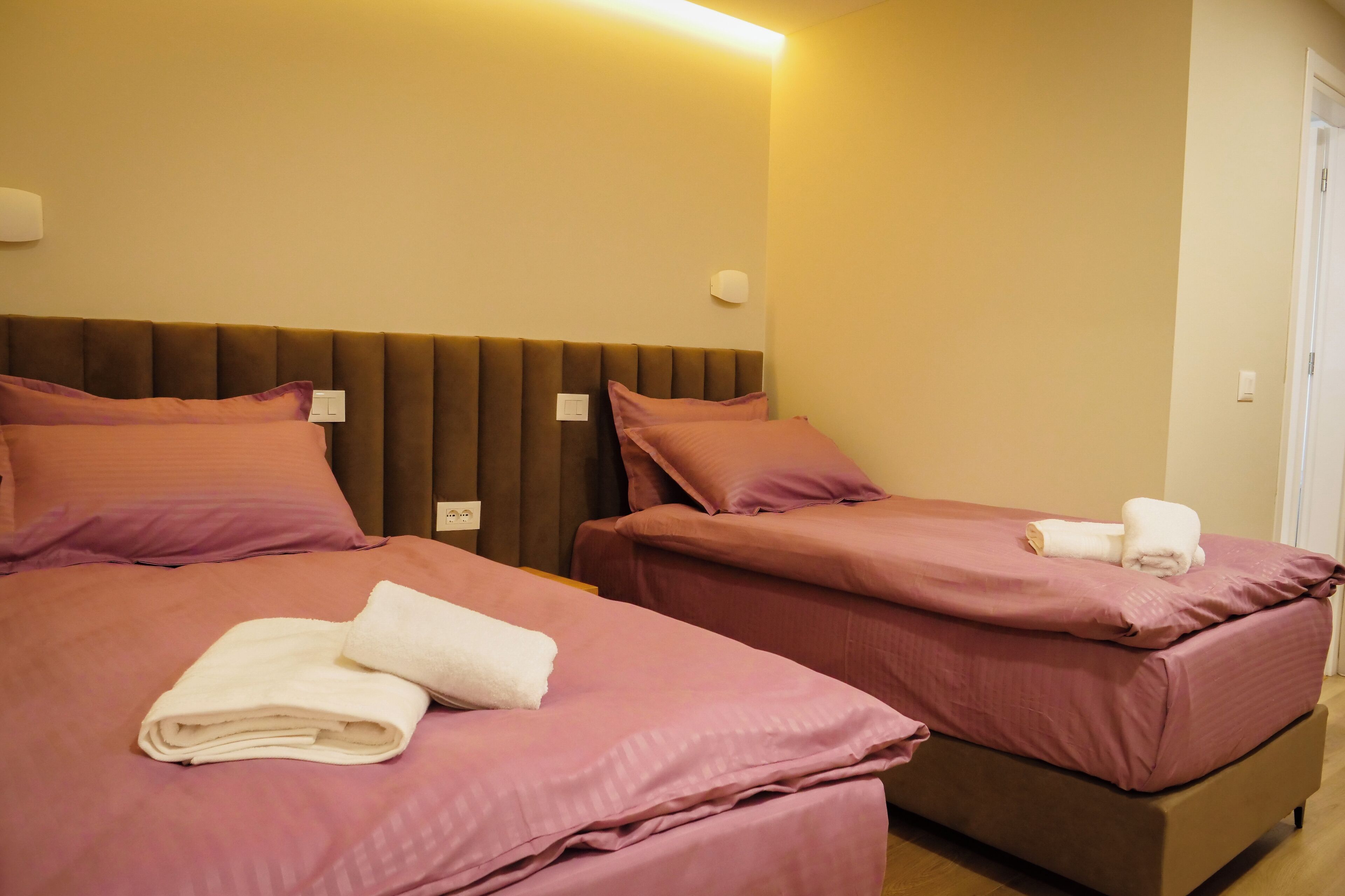 Dos camas con ropa de cama rosa y toallas blancas, cabecera marrón y lámpara fijada a la pared.