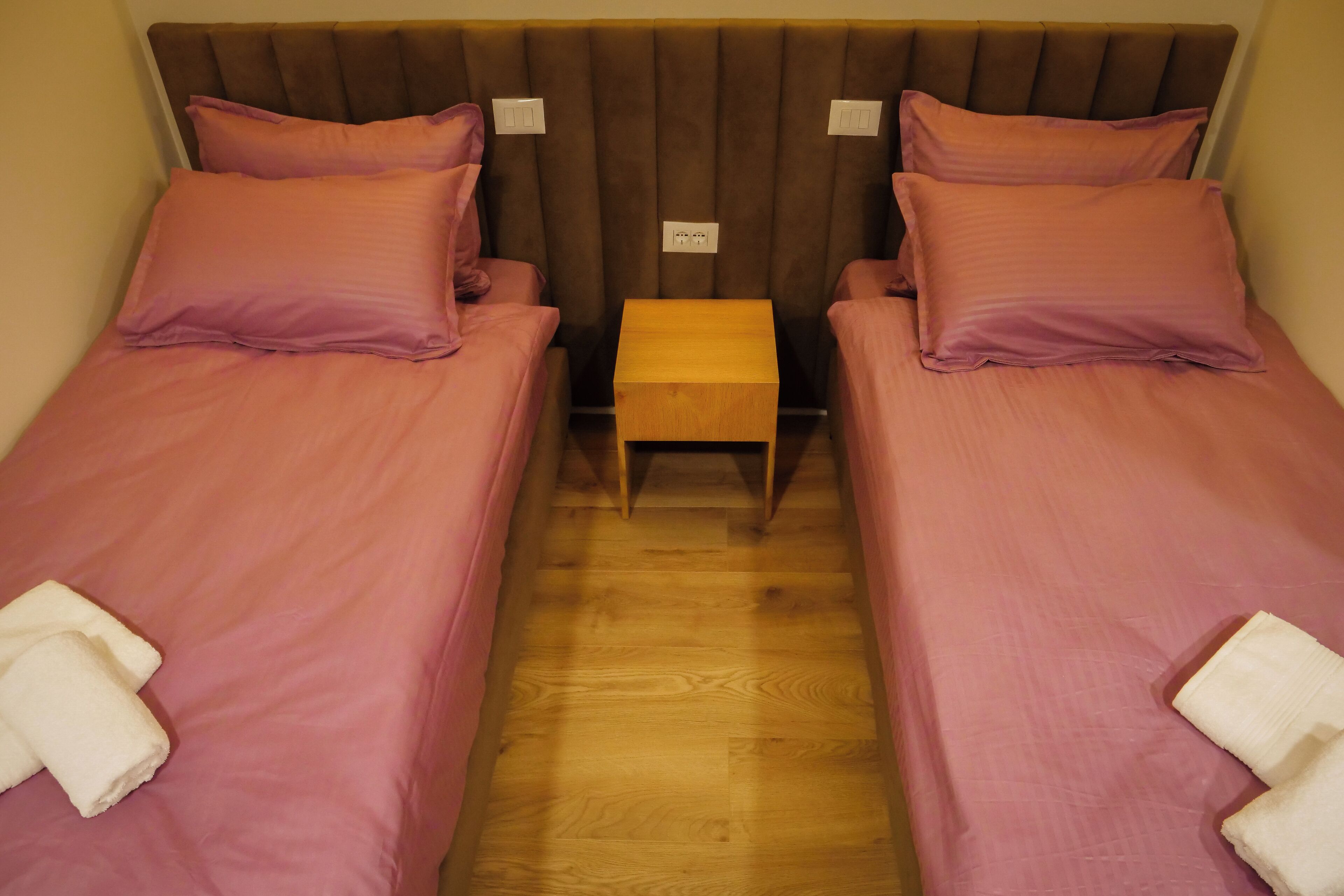 Una habitación con dos camas individuales, cada una con colcha y almohadas rosas a juego, una mesita de noche de madera entre ellas y un taburete de madera.