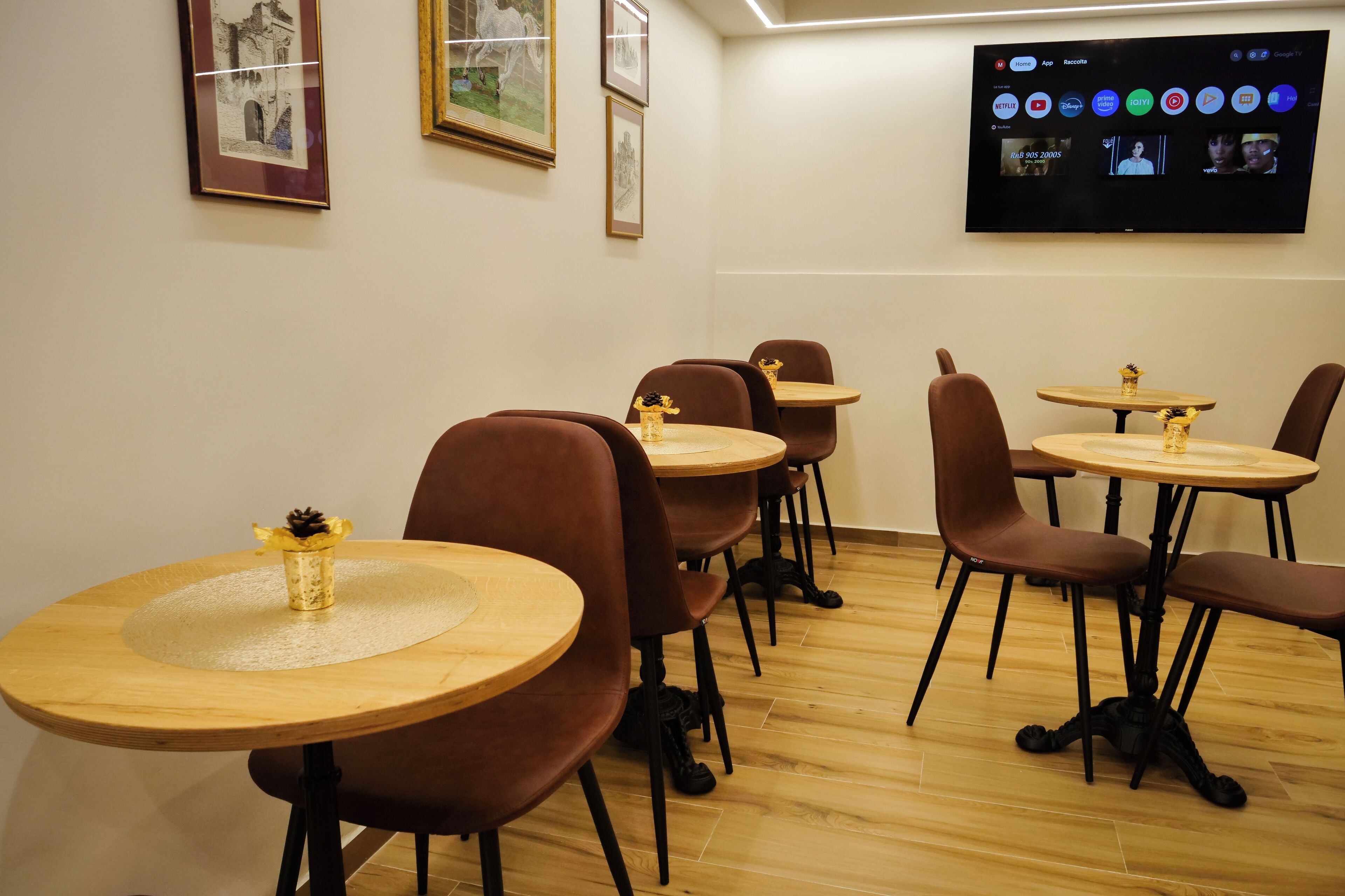 A conference room with multiple tables and chairs, a mounted television displaying various apps, and framed artwork on the walls.