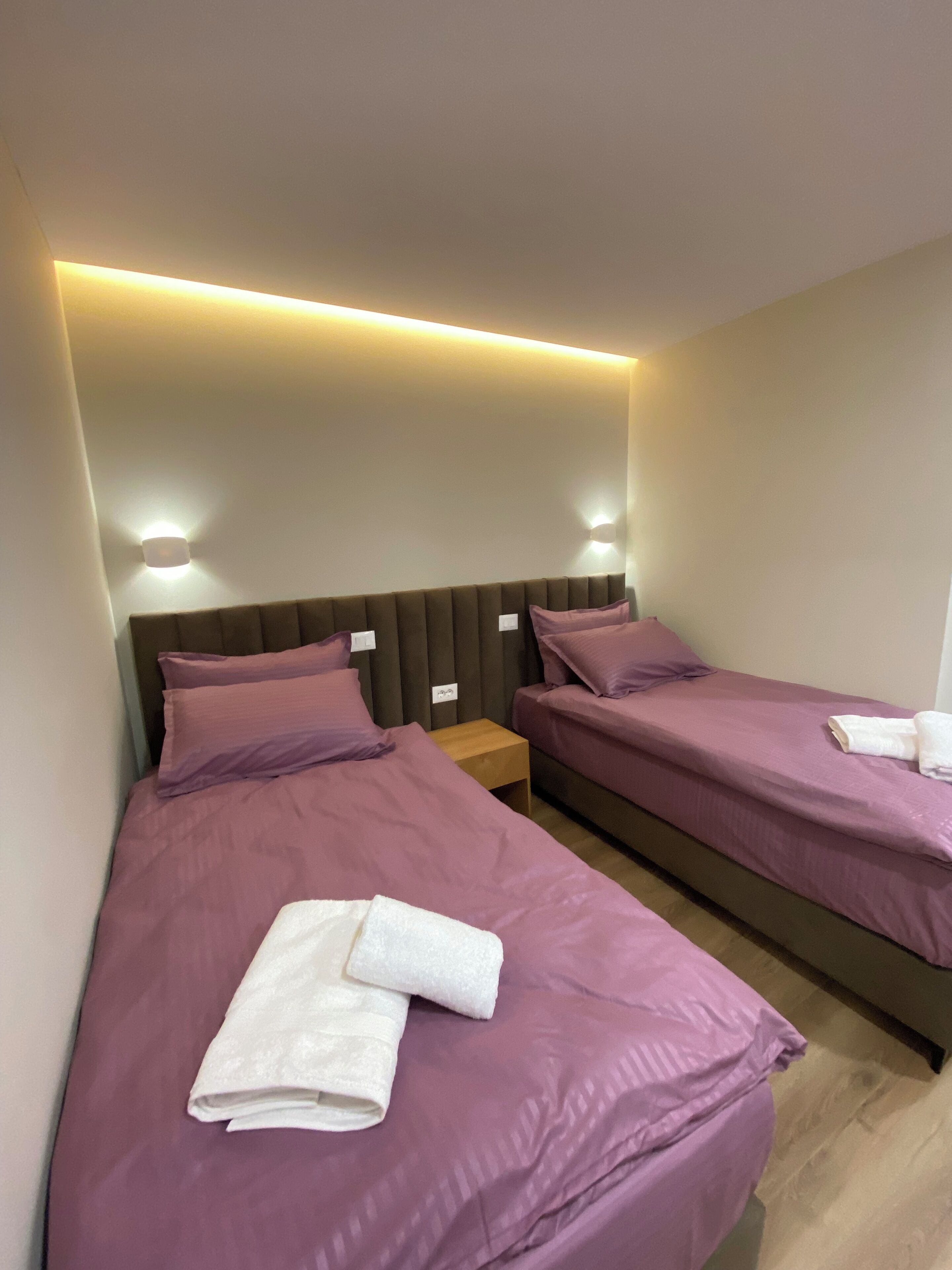 A hotel room with two beds, each with purple bedding and white towels. There are wall-mounted light fixtures above the beds.