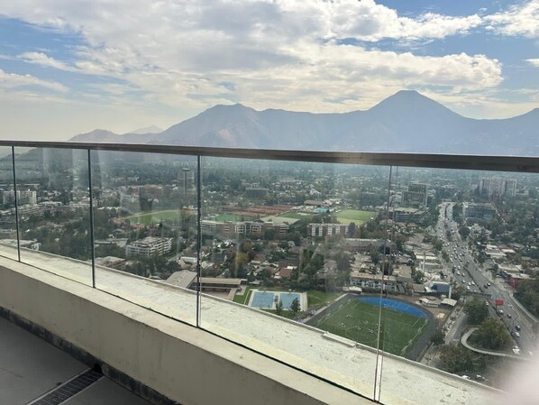 Un paisaje urbano con una cadena montañosa al fondo, visto desde un edificio de gran altura con barandilla de vidrio.