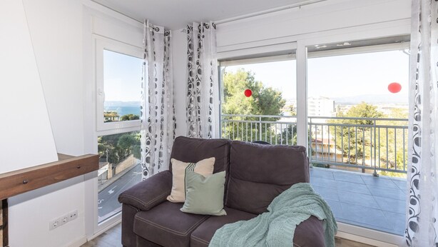 Ein modernes Wohnzimmer mit einer braunen Ledersofa, zwei Kissen und einer grünen Decke. Ein Holztisch steht neben dem Sofa. Das Zimmer verfügt über große Fenster mit gemusterten Vorhängen, welche einen Blick auf einen Balkon mit Geländer und einen roten Luftballon bieten.