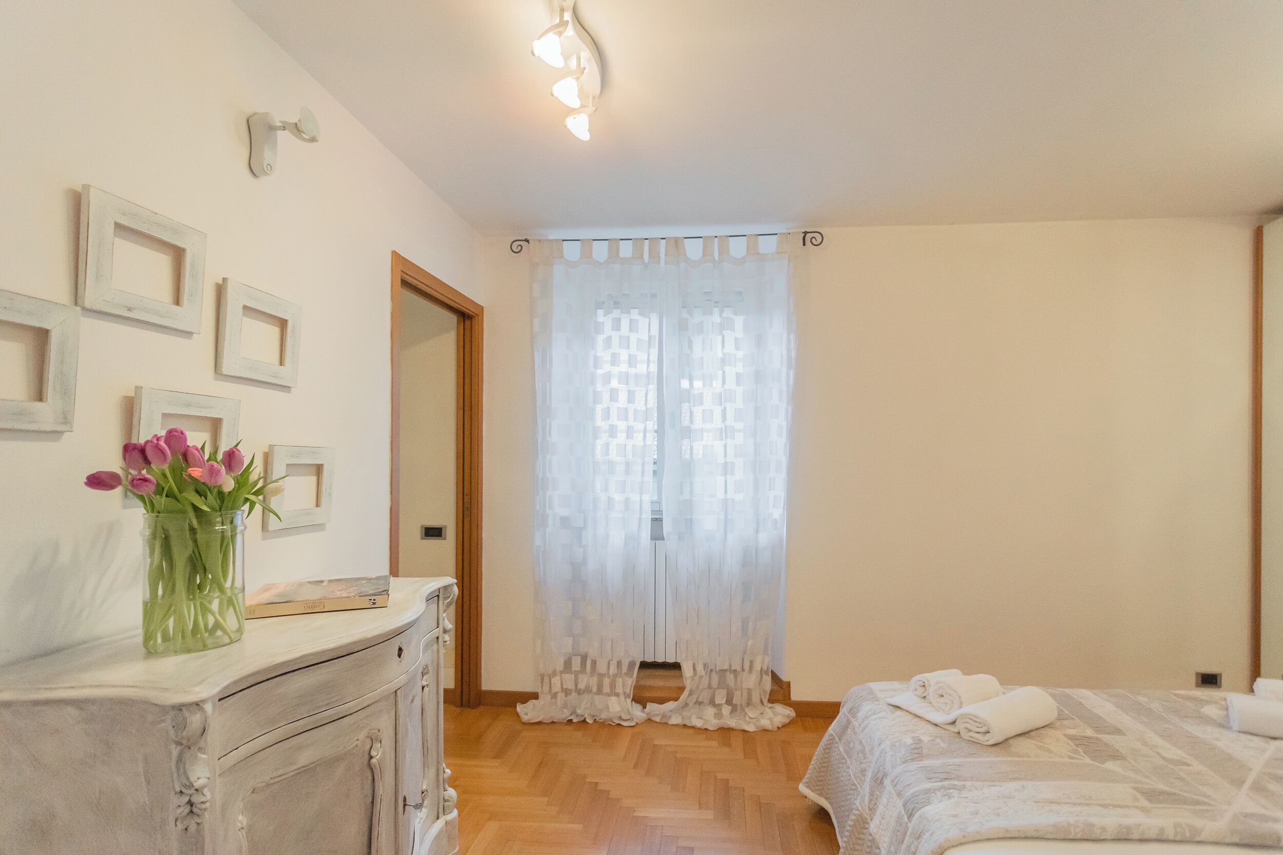 A bedroom with a wooden floor, a bed with white bedding, a dresser with a vase of flowers, and a window with sheer curtains.