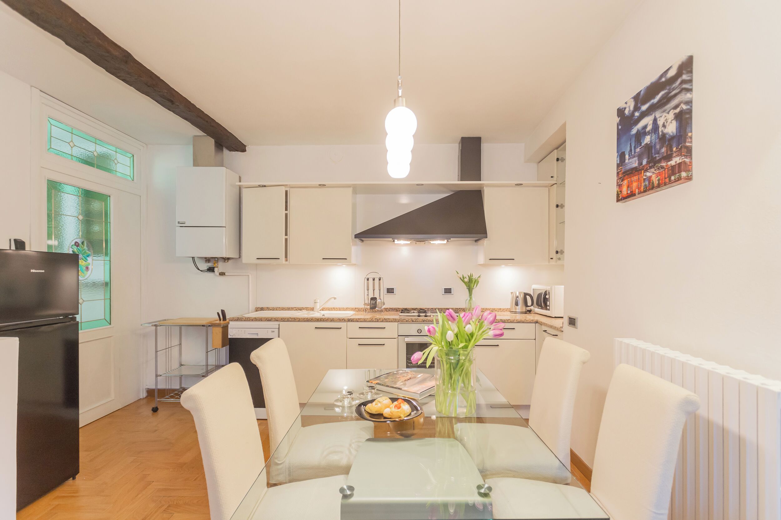 A modern kitchen with a glass dining table, white cabinets, and a cityscape painting.