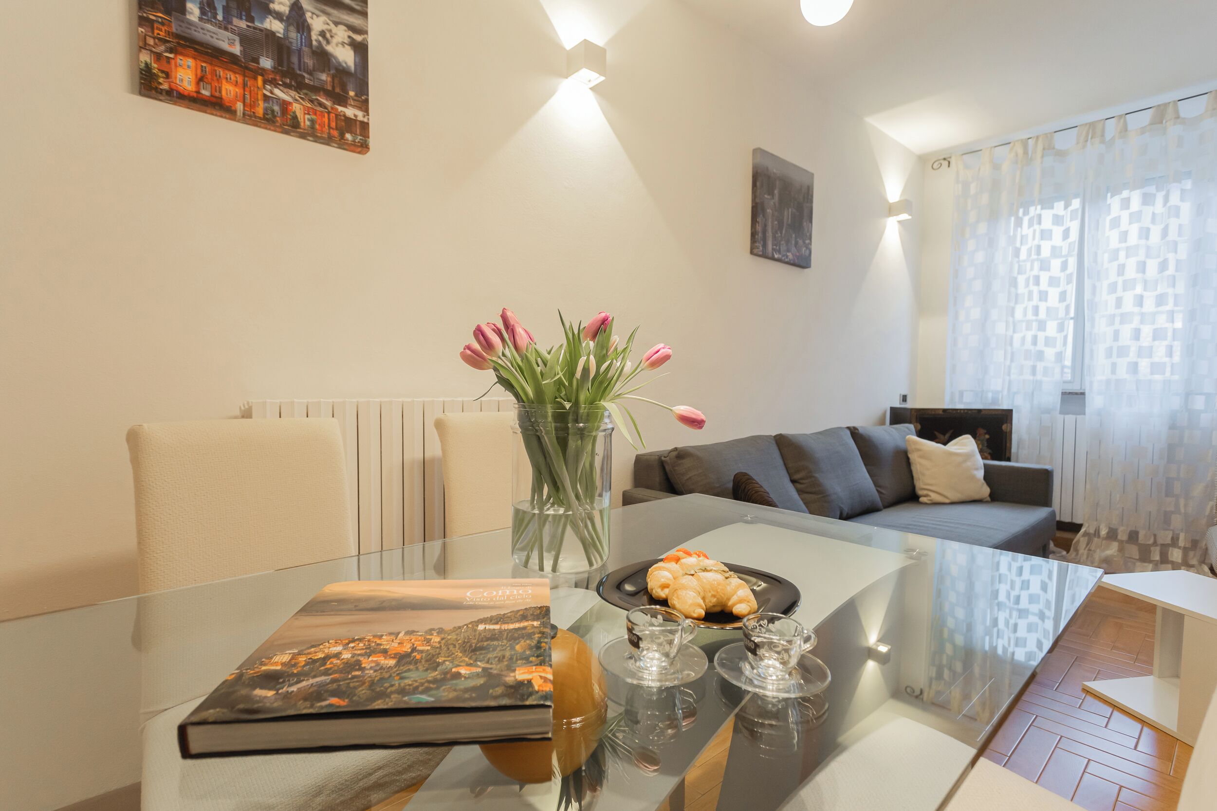 A modern living room with a glass dining table, a book, a vase with flowers, and a sofa with cushions.