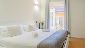 Una habitación de hotel con una cama grande, una ventana con cortinas, una mesita con un jarrón de flores y un balcón con barandilla.