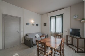 Een knus woongedeelte met een eettafel, stoelen, een bank, een televisie op een dressoir, en een deur.