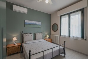 A bedroom with a bed, bedside tables, a radiator, a window with curtains, a wall-mounted air conditioner, and a painting.