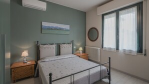 Un dormitorio con cama, mesitas de noche, un radiador, ventana con cortinas, aire acondicionado de pared y un cuadro.