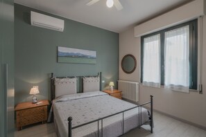 Un dormitorio con cama, mesitas de noche, un radiador, ventana con cortinas, aire acondicionado de pared y un cuadro.