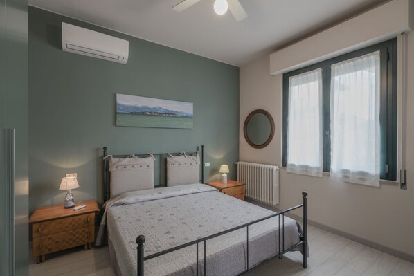 Une chambre à coucher comprenant un lit, des tables de chevet, un radiateur, une fenêtre avec des rideaux, un climatiseur mural et un tableau.