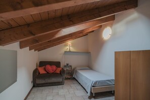 Een kleine, knusse kamer met een schuine plafond, een enkel bed, een bank met een rood kussen, een tafeltje met een lamp en een garderobe.