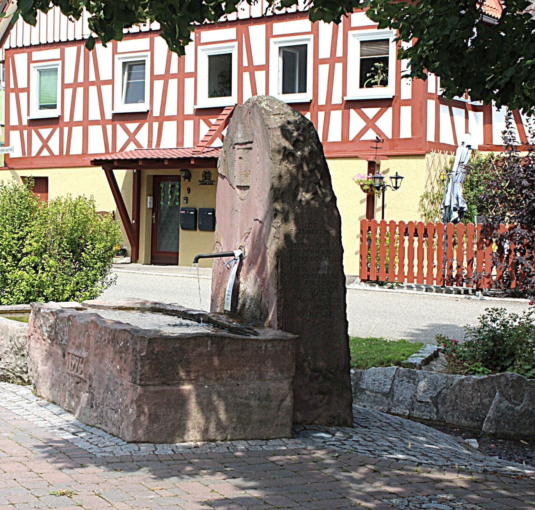 Ein Steinsockel mit Inschriften, ein Steinebecken und im Hintergrund ein Gebäude mit roter und weißer Fassade.