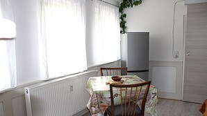 Un comedor con una mesa cubierta con mantel floral, sillas de madera, un cuenco en la mesa y un radiador debajo de la ventana.